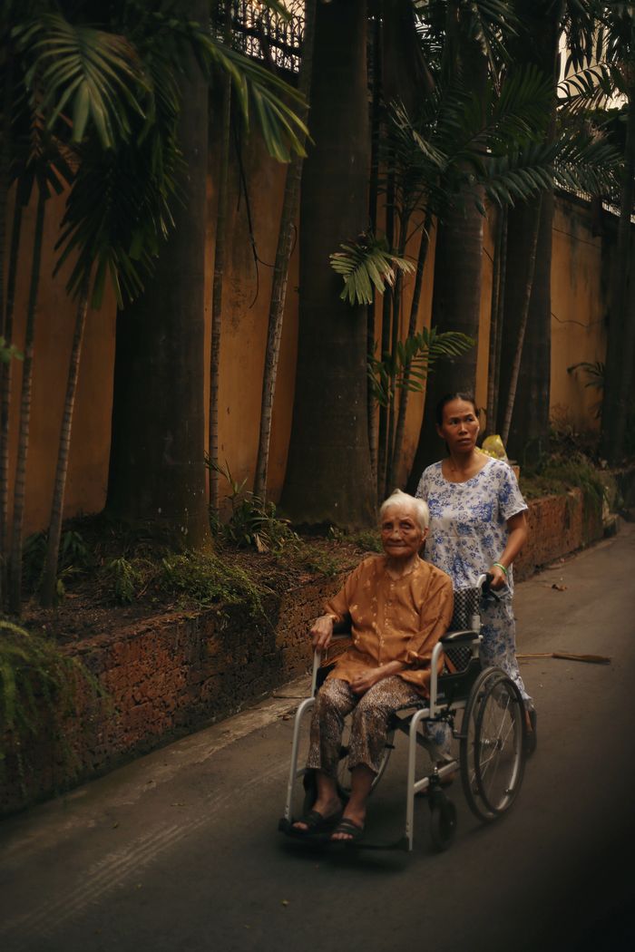 A caregiver pushing a senior woman in a wheelchair along a tropical street.