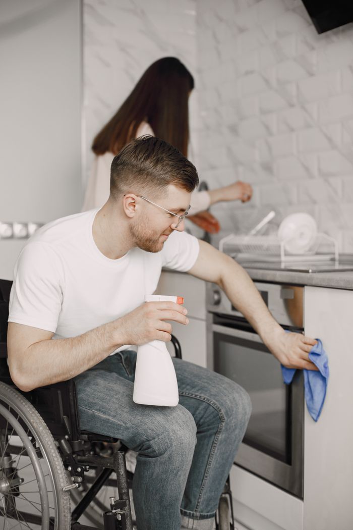 A man in a wheelchair cleaning the kitchen with spray and cloth, promoting inclusivity.